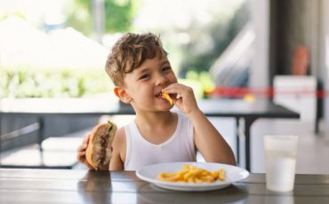 Little Boy Eating Sandwich and French Fries at Table. He appears focused on his meal, with a sandwich in one hand and a French Fries in the other hand. A boy eats fast food outdoors.